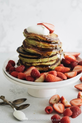 Close up of a stack of matcha pancakes with strawberry topping, whipped cream, and syrup